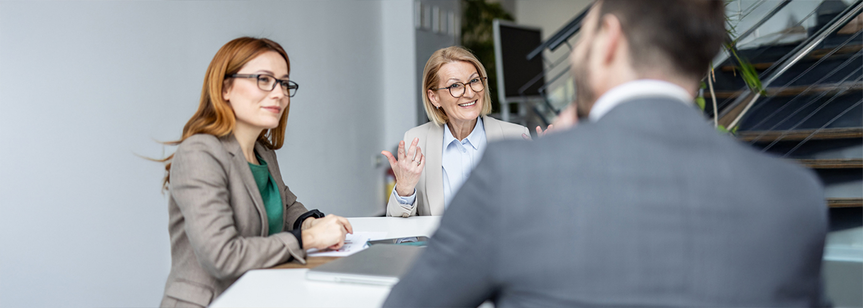 Zwei Frauen in Businesskleidung sitzen an einem Tisch und f&uuml;hren ein Gespr&auml;ch mit einem Mann im Anzug, der mit dem R&uuml;cken zur Kamera sitzt.