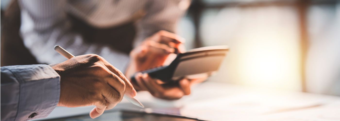 Zwei Personen am Schreibtisch. Die H&auml;nde sind sichtbar. Die Person im Vordergrund hat einen Stift in der Hand, die Person  im Hintergrund einen Taschenrechner.