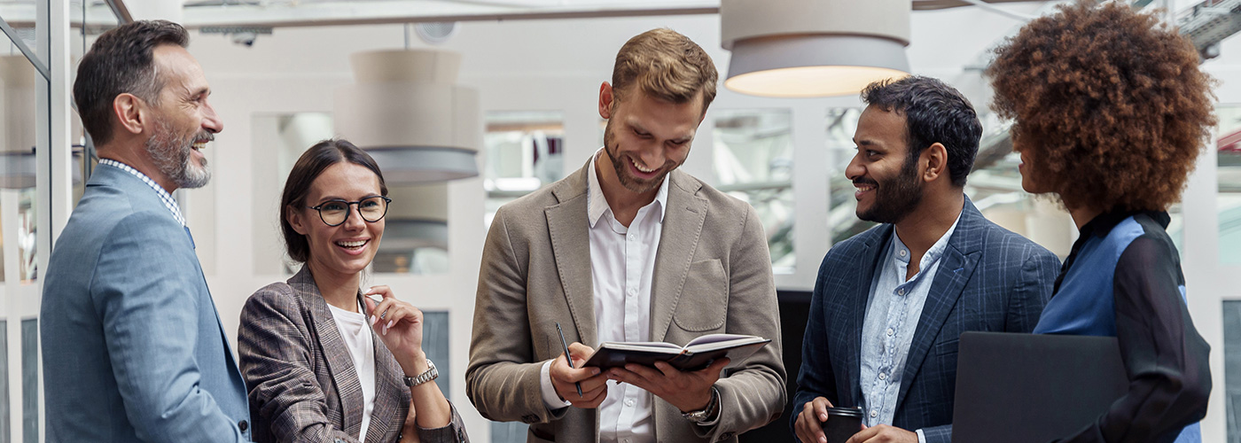 Ein dynamisches Team von fünf Geschäftsleuten tauscht sich in einer modernen Büroumgebung aus. Die lockere und freundliche Atmosphäre, begleitet von Lachen und positiver Energie, unterstreicht die Bedeutung von Teamarbeit, Kommunikation und beruflicher Entwicklung.