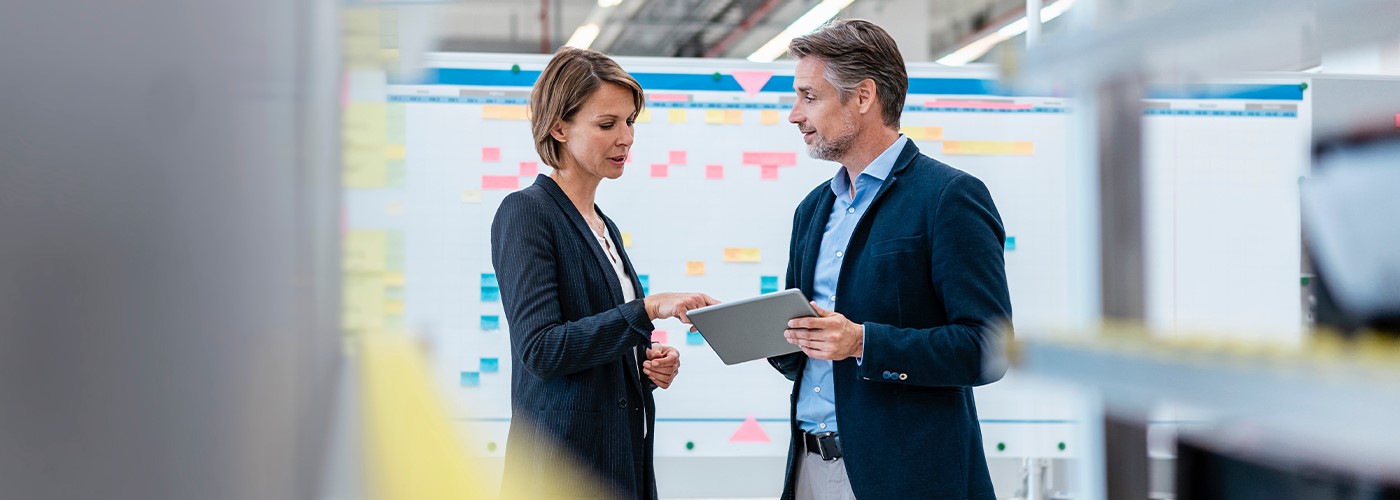 Zwei Personen im Gespräch vor einem Whiteboard mit bunten Notizen, betrachten gemeinsam ein Tablet in einer modernen Arbeitsumgebung.