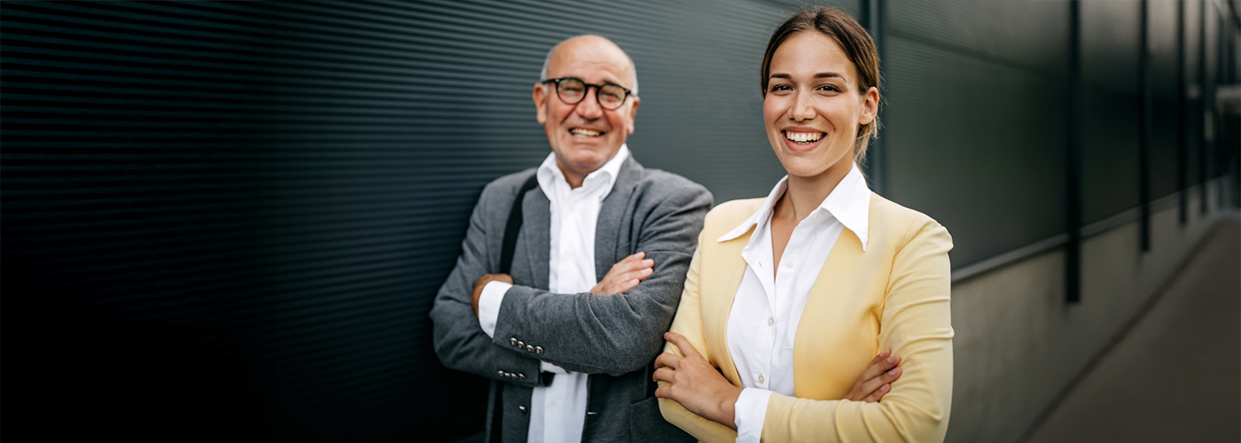 Zwei l&auml;chelnde Personen, ein &auml;lterer Mann mit Brille und grauem Sakko und eine j&uuml;ngere Frau mit gelbem Blazer, stehen vor einer dunklen Wand mit verschr&auml;nkten Armen.