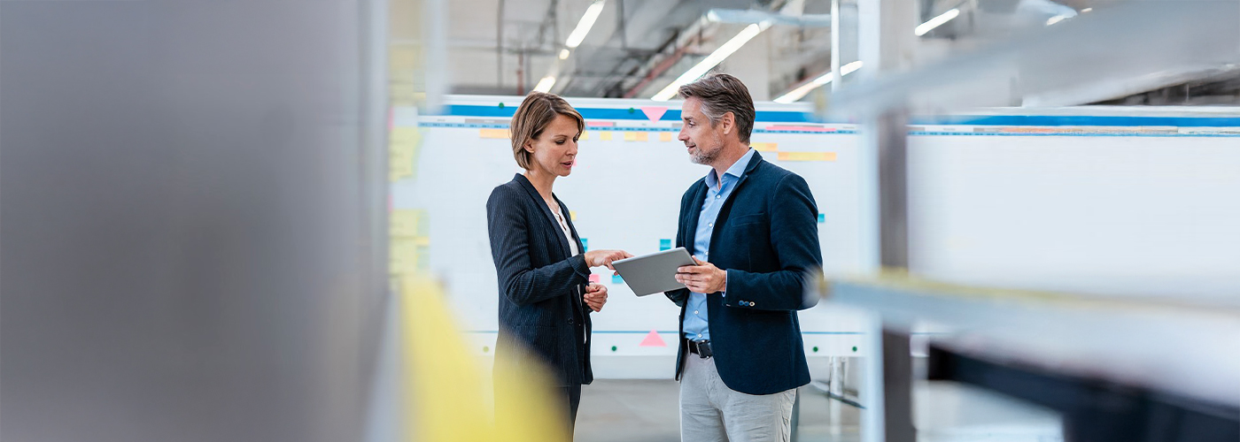 Two people in conversation in front of a whiteboard with colourful notes, looking together at a tablet in a modern working environment.