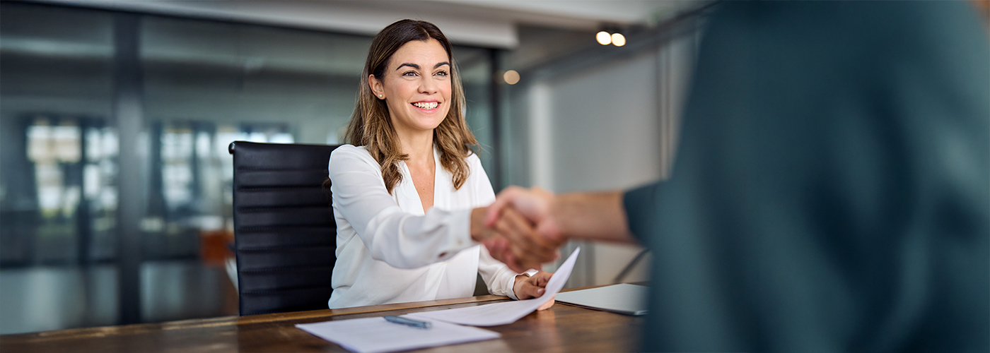 A smiling woman sits at a desk in an office and shakes a person's hand.