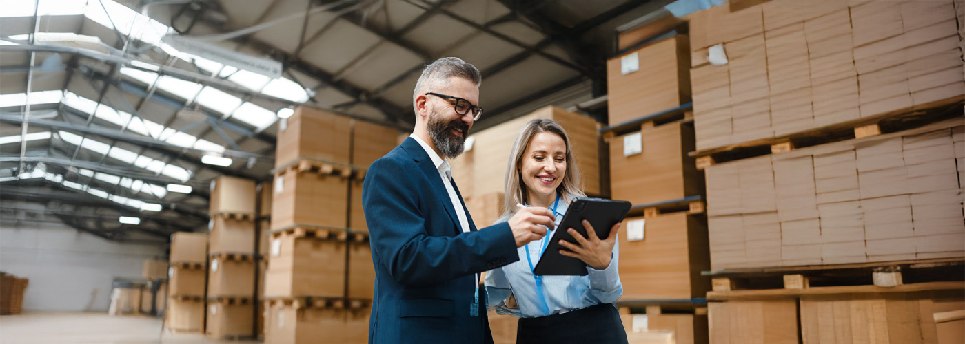Ein Mann und eine Frau in einer Lagerhalle mit gestapelten Paletten im Hintergrund.