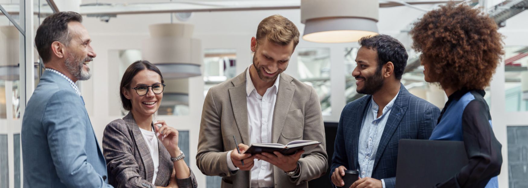 Ein dynamisches Team von f&uuml;nf Gesch&auml;ftsleuten tauscht sich in einer modernen B&uuml;roumgebung aus. Die lockere und freundliche Atmosph&auml;re, begleitet von Lachen und positiver Energie, unterstreicht die Bedeutung von Teamarbeit, Kommunikation und beruflicher Entwicklung.