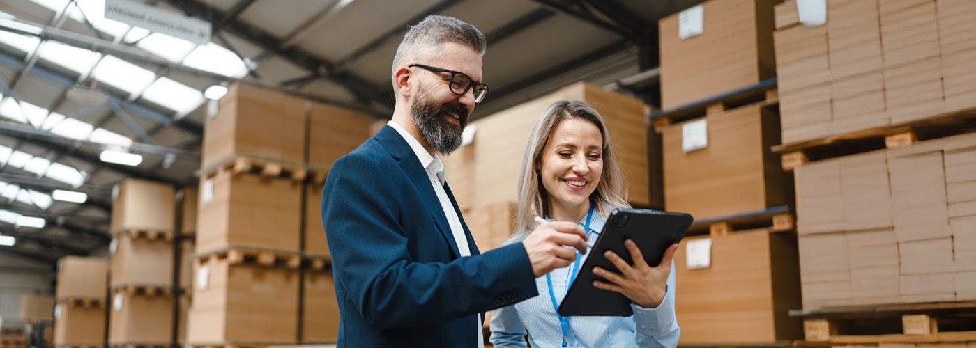 A man and a woman in a warehouse with packed, stacked pallets in the background. The woman is holding a clipboard, the man a pen. Both are looking at the clipboard.