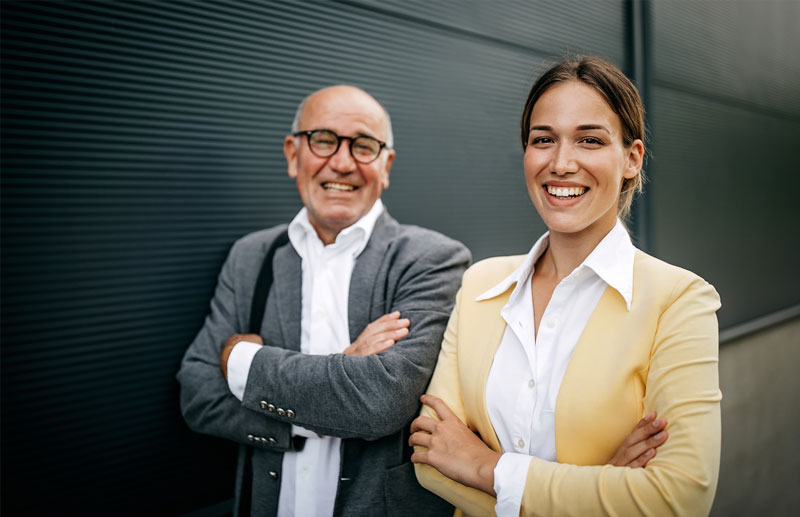 Zwei l&auml;chelnde Personen, ein &auml;lterer Mann mit Brille und grauem Sakko und eine j&uuml;ngere Frau mit gelbem Blazer, stehen vor einer dunklen Wand mit verschr&auml;nkten Armen.