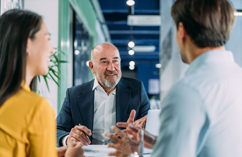 Drei Personen sitzen an einem Besprechungstisch in einem B&uuml;ro und unterhalten sich &uuml;ber eine Firmen&uuml;bergabe.