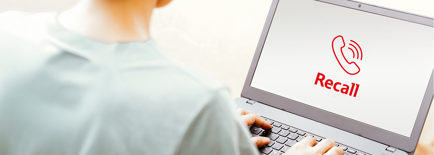 A person is sitting at a laptop with their back to the viewer. An recall pictogram in red with the text recall can be seen on the screen.