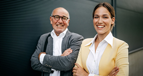 Factoring in der Unternehmensnachfolge Zwei lächelnde Personen, ein älterer Mann mit Brille und grauem Sakko und eine jüngere Frau mit gelbem Blazer, stehen vor einer dunklen Wand mit verschränkten Armen.