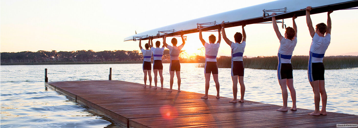 Eine Gruppe eines Ruderclubs tr&auml;gt gemeinsam ein Kajak &uuml;ber den K&ouml;pfen. Es symbolisiert, dass alle im selben Boot sitzen.