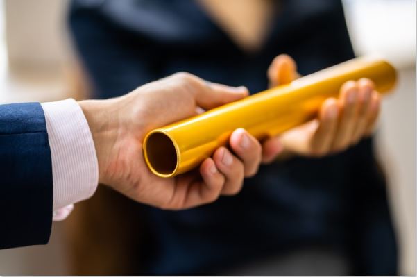 Zwei Personen in Businesskleidung &uuml;bergeben sich eine goldfarbene Urkundenrolle im Vordergrund