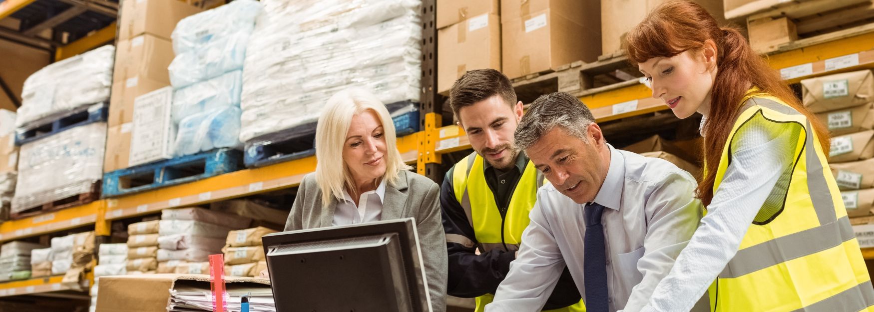 Four people are standing in a warehouse, looking cheerfully at a computer screen together.