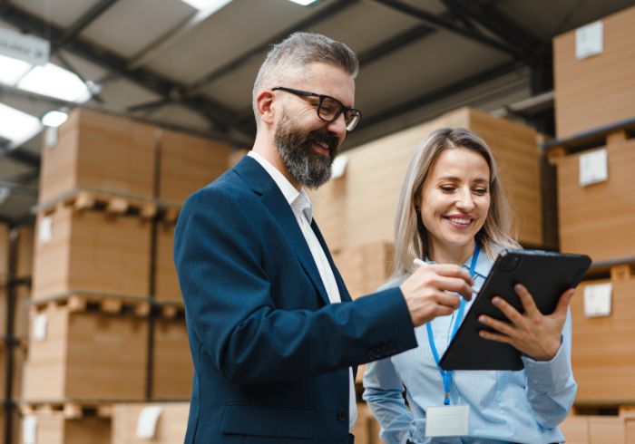 A man and a woman in a warehouse with packed, stacked pallets in the background. The woman is holding a clipboard, the man a pen. Both are looking at the clipboard.