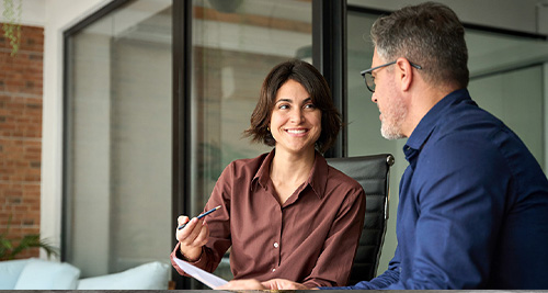 Zwei Personen sitzen an einem Tisch mit Laptop und Dokumenten in einem modernen B&uuml;ro.