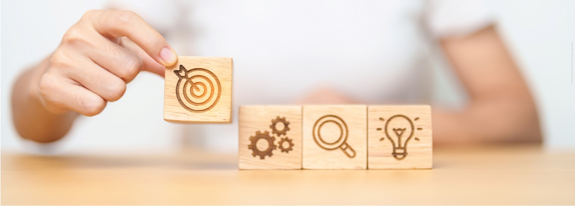 One hand places a wooden block with a target as a symbol for goal achievement on a series of other blocks. These show cogwheels (processes), a magnifying glass (analysis) and a light bulb (idea).