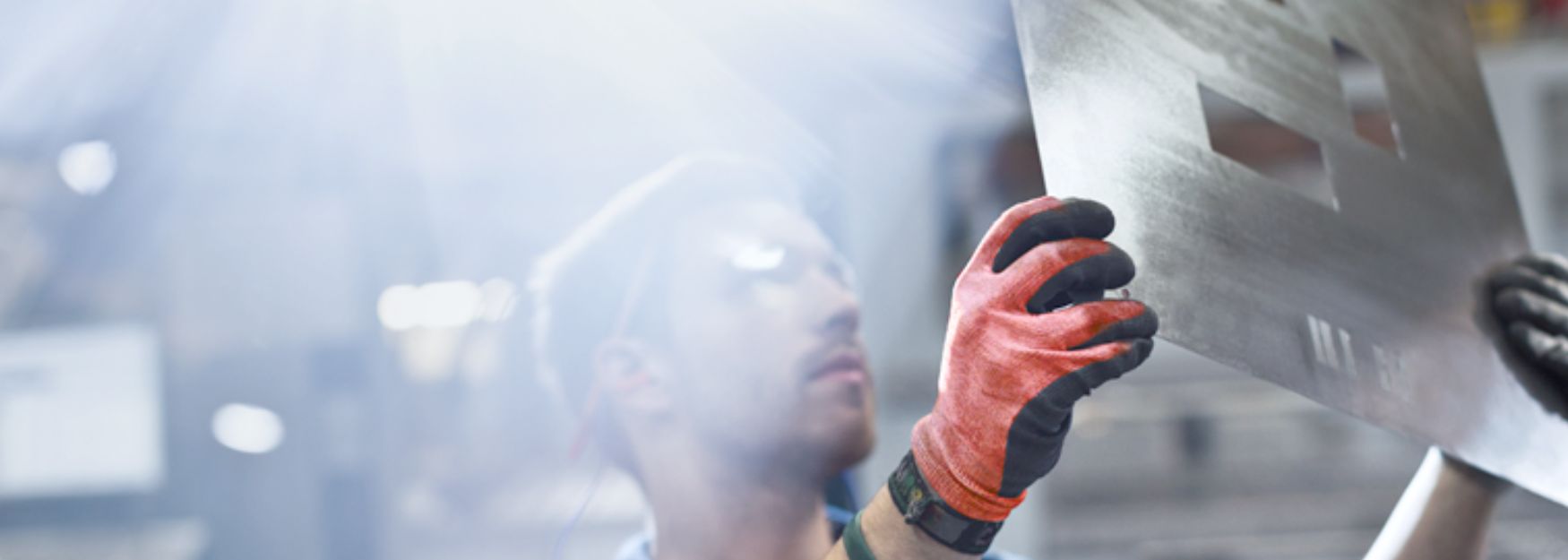 Ein Mann hält eine Metallplatte in einer Produktionshalle mit roten Schutzhandschuhen und begutachtet diese.